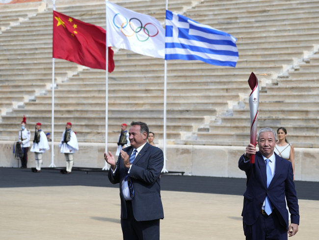 Snímek: Viceprezident BOCOG Yu Zaiqing (Jü Caj-čching, vpravo) zvedá pochodeň, zatímco prezident Řeckého olympijského výboru Spyros Capralos aplauduje během ceremoniálu předávání olympijského ohně v Athénách, Řecko; 19. října 2021 / CFP